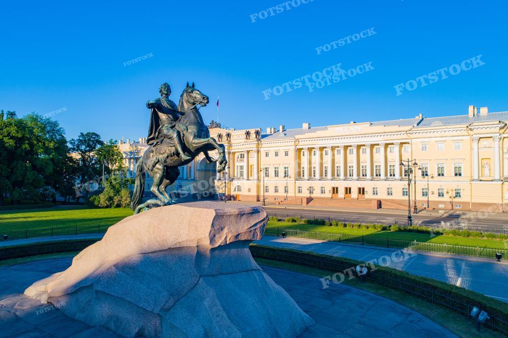 На фотографии изображён Медный всадник — знаменитый памятник Петру I в Санкт-Петербурге, расположенный на Сенатской площади. Фоном для монумента служат зелёные насаждения парка и здание Сената и Синода в лучах утреннего солнца. Светло-голубое небо придаёт изображению спокойствие и свежесть. Фотографию можно использовать в качестве иллюстрации для исторических и культурных материалов, туристических путеводителей и образовательных ресурсов.